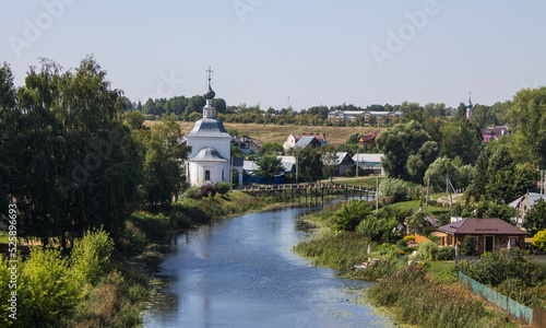 The white stone Church of Elijah the Prophet on the bank of the Kamenka river in Suzdal Russia on a summer day and old houses among lush green foliage