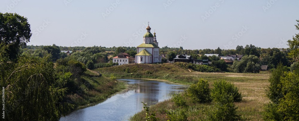 The white stone Church of Elijah the Prophet on the bank of the Kamenka river in Suzdal Russia on a summer day and old houses among lush green foliage