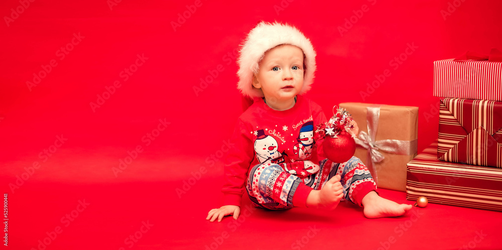 Little Santa with a Christmas tree toy in his hands sits next to Christmas presents on a red background Little Santa with a Christmas tree toy in his hands sits next to Christmas presents on a red background