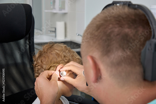 View from above examination by otolaryngologist of eardrum of the child's ear with the help of the ear funnel. Curly boy sits in a chair at the reception at the ENT, checks his ears for traffic jams