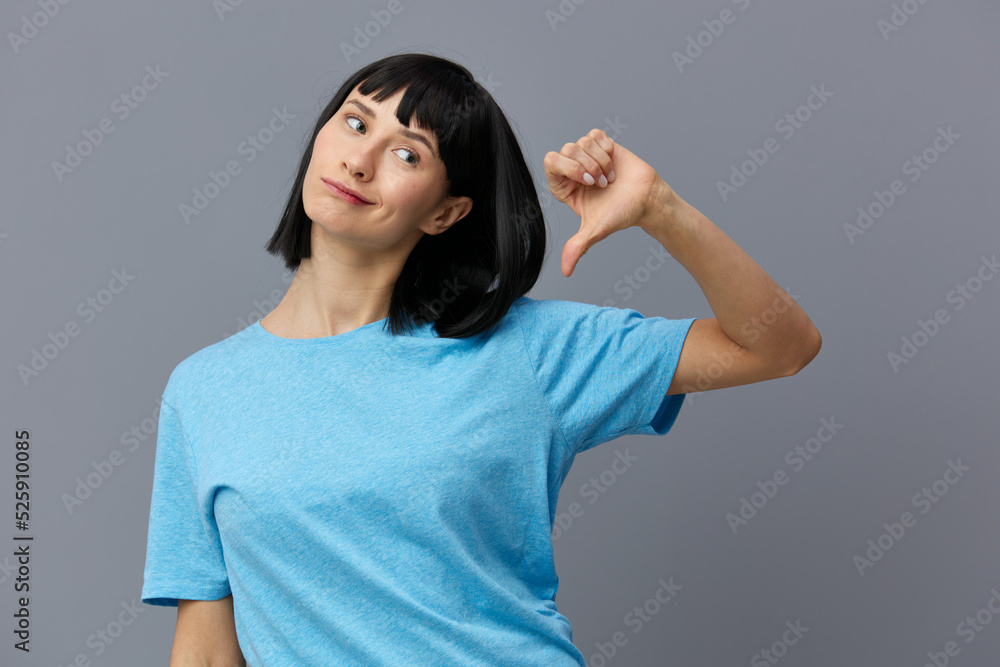a lovely woman stands on a gray background in a blue T-shirt, happily grinning, raises her hand up showing a dislike a lovely woman stands on a gray background in a blue T-shirt, happily grinning, raises her hand up showing a dislike