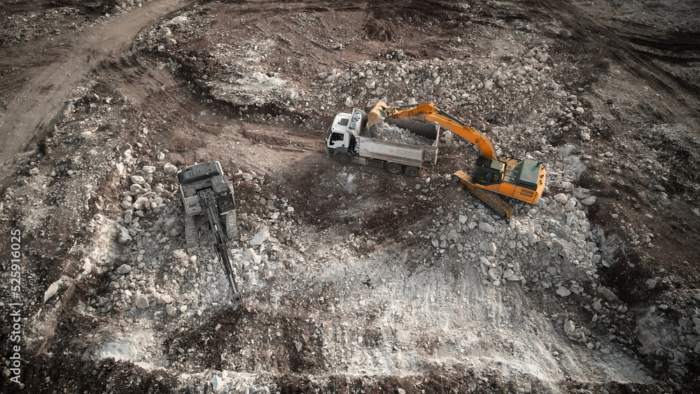 Obraz premium Aerial view of excavator loading tipper truck with crushed rock soil. Machineries at construction site. Earthworks. Heavy machinery at earthmoving works