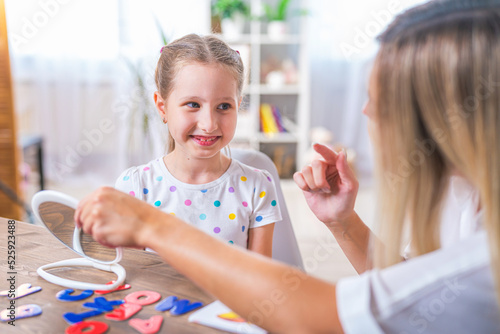 Doctor and small patient train articulation, work on problems and obstacles child with dyslexia. little girl together with speech therapist is sitting at desk indoors, playing game, studying sounds photo