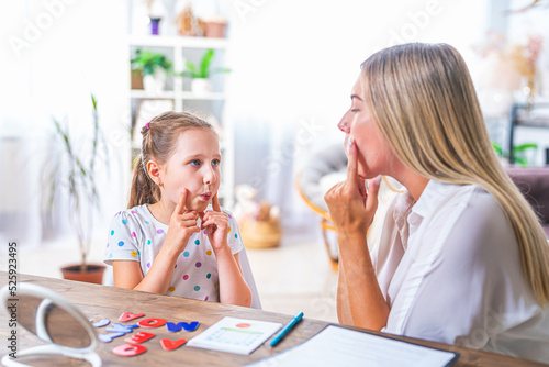 Doctor and small patient train articulation, work on problems and obstacles child with dyslexia. little girl together with speech therapist is sitting at desk indoors, playing game, studying sounds