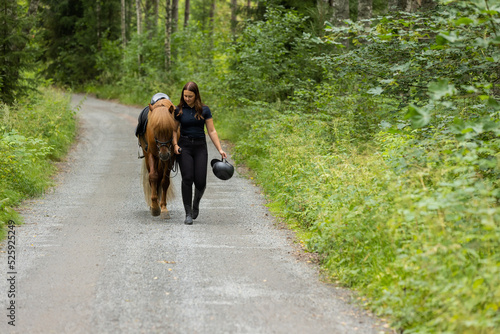 Icelandic horse with female rider. Horse and rrider walk side by side. Rider carries helmet in hand.