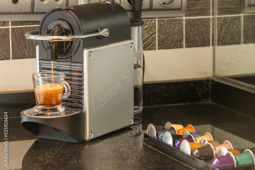 Capsule coffee machine on a black countertop, and a transparent cup filling with coffee and capsules of various kinds