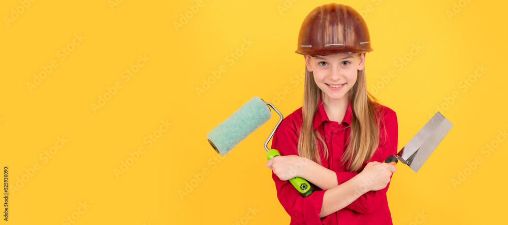 Fototapeta premium positive teen child in builder hard hat with paint roller and spatula on yellow wall. Child builder in helmet horizontal poster design. Banner header, copy space.