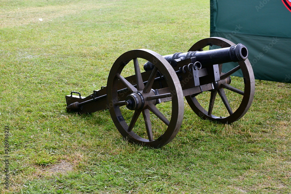 A close up on a replica of a Napoleonian cannon made out of metal and wood standing in the middle of a field or meadow next to a green tent seen during a hike on a Polish countryside