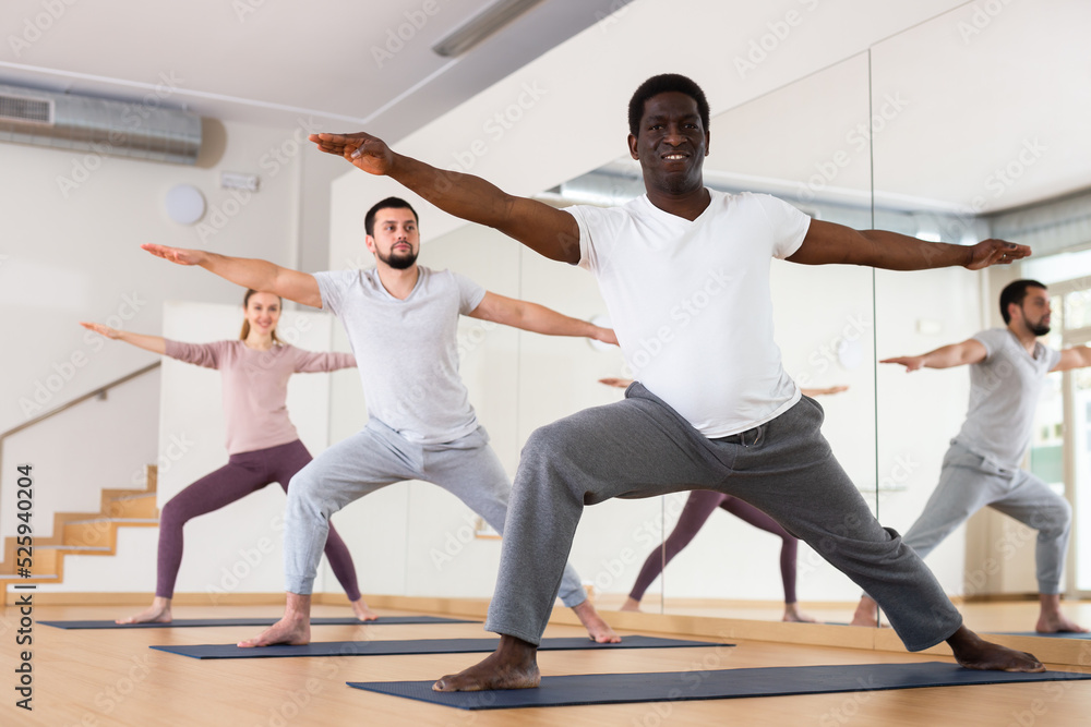 Sporty adult African American doing power yoga with group in fitness studio, standing in lunging asana Virabhadrasana..