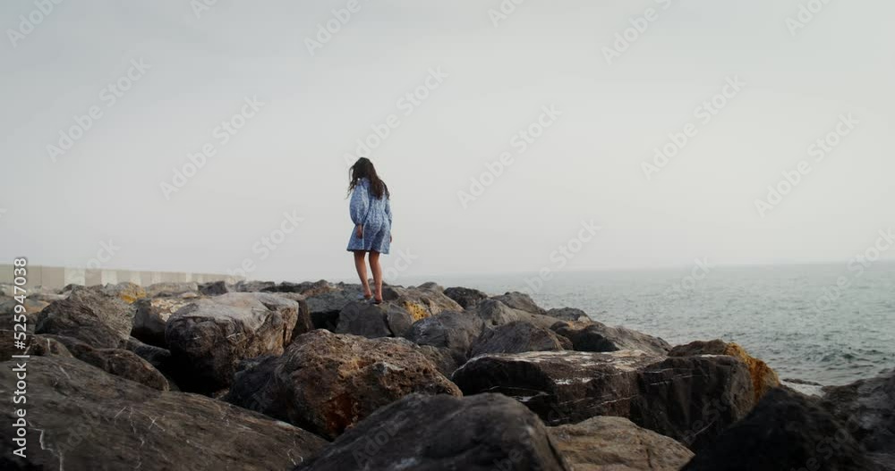 A young woman with flowing curly dark hair walks over large boulders, stepping from stone to stone, along the coastline on a clear summer day. 4k video, red komodo A young woman with flowing curly dark hair walks over large boulders, stepping from stone to stone, along the coastline on a clear summer day. 4k video, red komodo