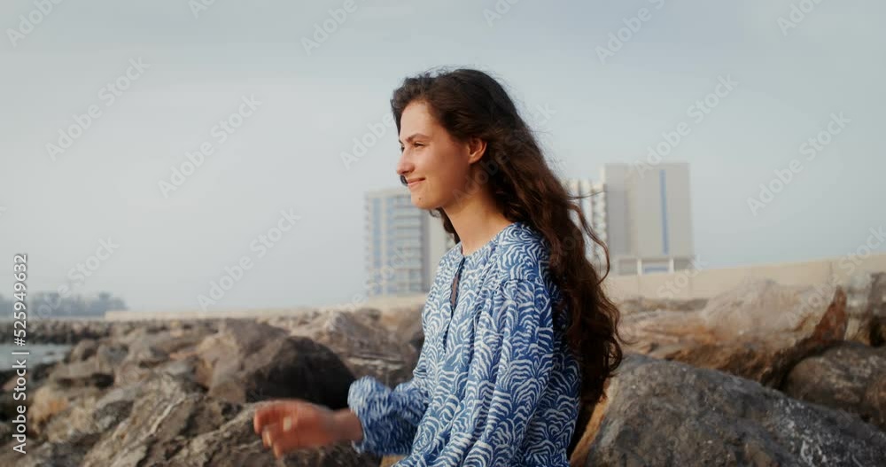 A beautiful young woman smiles and straightens her hair looking into the distance, sitting on large stones on the seashore. 4k video, red komodo A beautiful young woman smiles and straightens her hair looking into the distance, sitting on large stones on the seashore. 4k video, red komodo