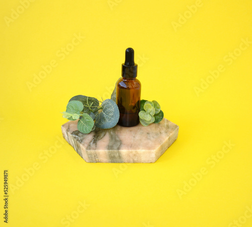 Glass dropper bottle with black lid on a podium with  plants on yellow background. Beauty concept for face and body care.