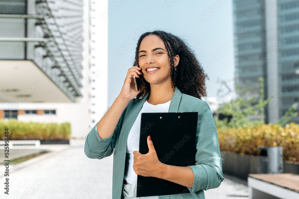 Cell Phone conversation. Confident positive young mixed race curly successful business woman, real estate agent, in elegant clothes, has conversation on a smartphone while standing outdoors, smiling Cell Phone conversation. Confident positive young mixed race curly successful business woman, real estate agent, in elegant clothes, has conversation on a smartphone while standing outdoors, smiling