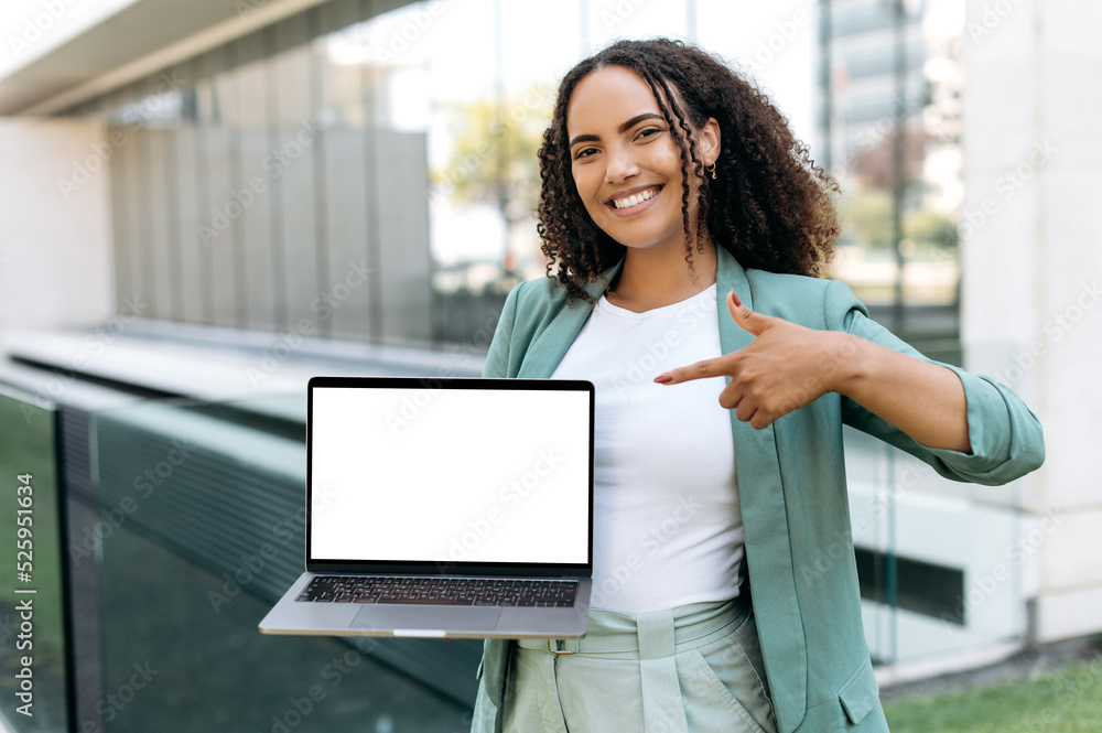 Positive lovely successful young woman with curly hair, stylishly dressed, stands outdoors, holds an open laptop with blank white mock-up screen in hand, points finger at it, looks at camera, smiles Positive lovely successful young woman with curly hair, stylishly dressed, stands outdoors, holds an open laptop with blank white mock-up screen in hand, points finger at it, looks at camera, smiles