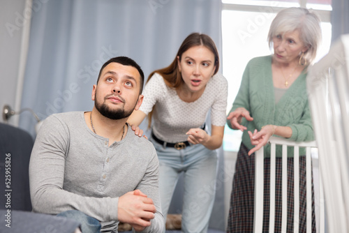 Offended man sitting on sofa in apartment. His wife and mother-in-law quarreling with him.