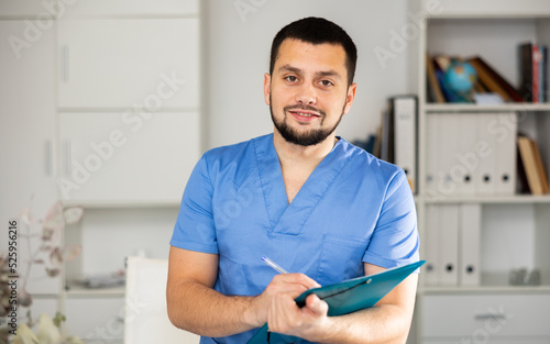 Positive man physician standing in his office  holding paper folder and looking at camera.