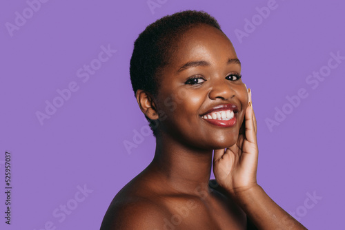 Portrait of attractive African girl touching clean cheeks with perfect glow, isolated on purple background with free space for text. Smiling and looking at