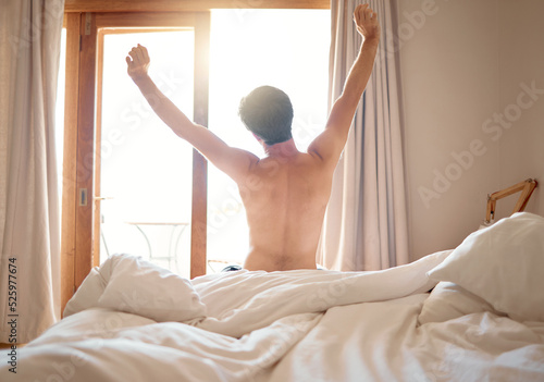 Man in nude stretching after night sleep in bed, waking up to morning sunrise in bedroom and feeling excited for day at home. Back of naked male person relaxing after nap or rest in apartment house