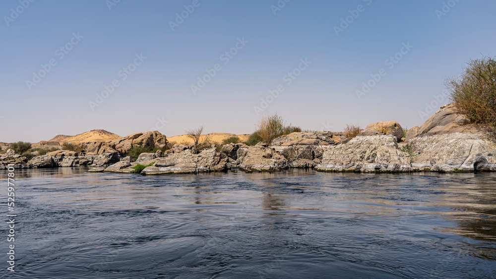 A blue calm river with rocky shores. Picturesque boulders and sand dunes against a clear sky. Ripples and reflections on the water. Copy space. Egypt. Nile