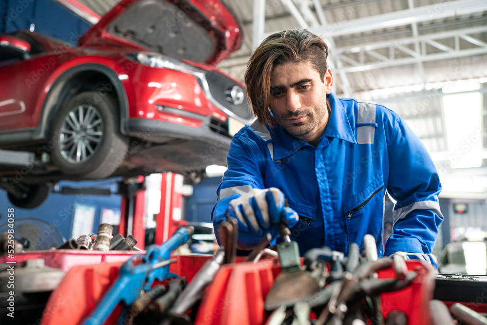 Male car operator wearing blue overalls, gloves working under the hood of car and checking attentively serviceability of engine at repair garage. Concept of car maintenance