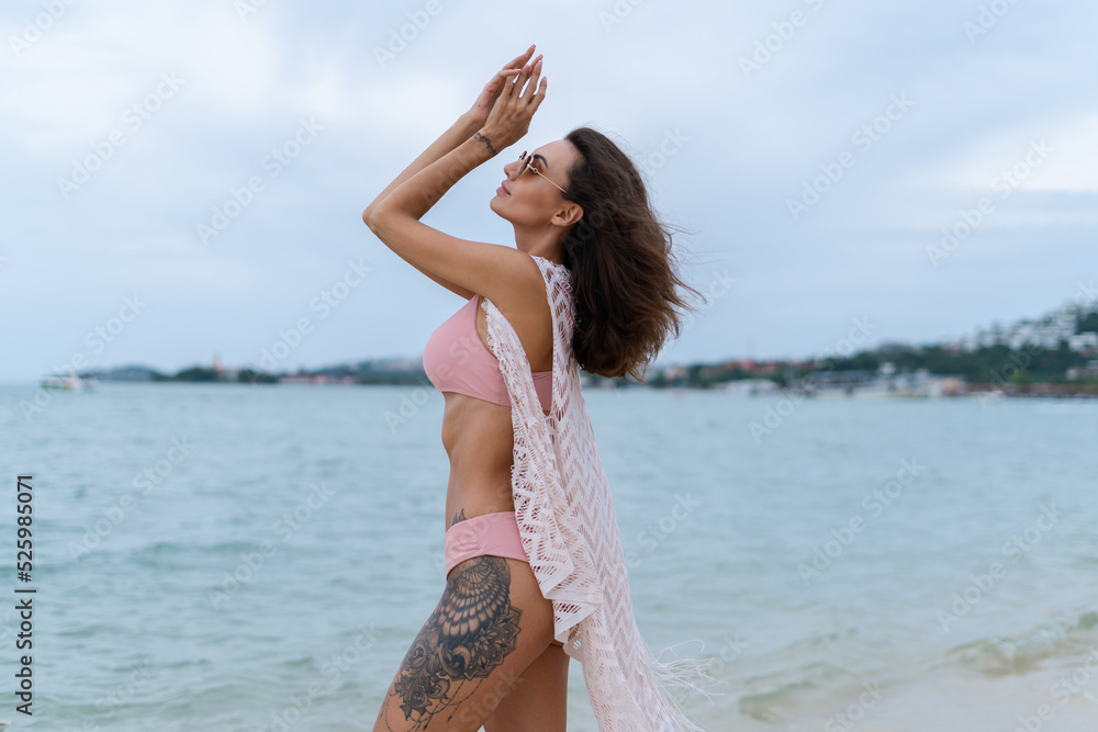 Naklejka premium Stylish young woman with voluminous hair in a bikini and lace pareo posing on a tropical beach, slim, tanned, athletic, sunglasses