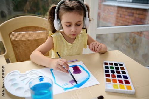 Adorable Caucasian preschooler girl in wireless headphones, concentrated little artist, drawing picture, using watercolor paints. Kids entertainment, hobby and education concept. Art master class.