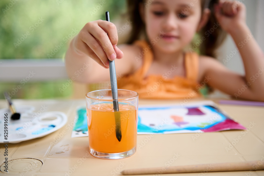 Selective focus on a glass with colored water and hand of blurred Caucasian child, preschooler girl dipping a paintbrush while drawing and painting with watercolors. Creativity. Creative art courses Selective focus on a glass with colored water and hand of blurred Caucasian child, preschooler girl dipping a paintbrush while drawing and painting with watercolors. Creativity. Creative art courses