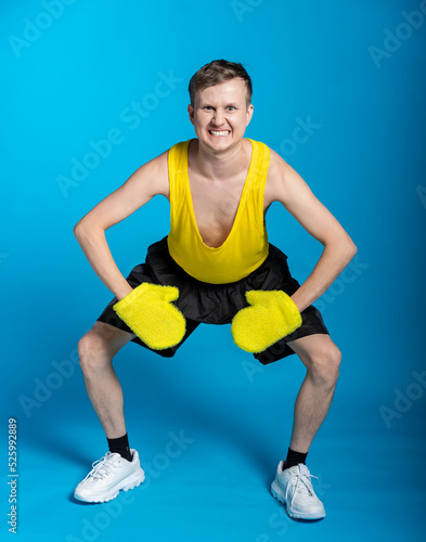 lean sportsman in a yellow t-shirt and black shorts on a blue background. funny jock put on yellow massage mittens and funny grimaces face