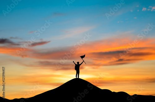 Silhouette of young businessman standing alone on top of the mountain and hand holding flag looking beautiful view sunrise. He raised both arms, showing joy and success.