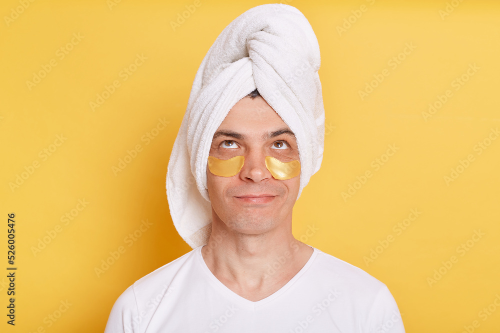 Portrait of pensive confused man wearing white t shirt and towel, having cosmetic patches under his eyes, looking up with puzzled facial expression, posing isolated over yellow background. Portrait of pensive confused man wearing white t shirt and towel, having cosmetic patches under his eyes, looking up with puzzled facial expression, posing isolated over yellow background.