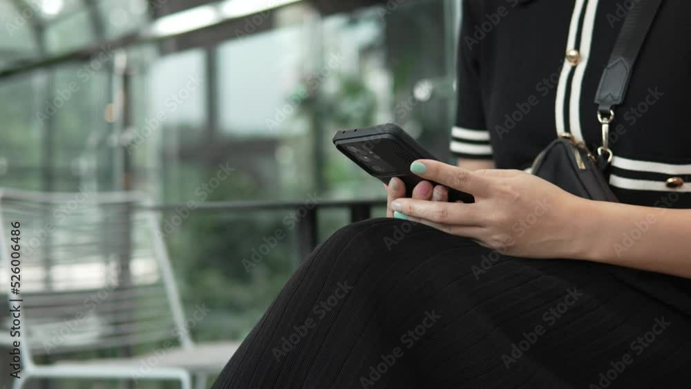 Young woman dressed in black dress uses the phone while sitting on a chair in a modern shopping mall, close-up of hands with phone, the woman's face is not visible Young woman dressed in black dress uses the phone while sitting on a chair in a modern shopping mall, close-up of hands with phone, the woman's face is not visible