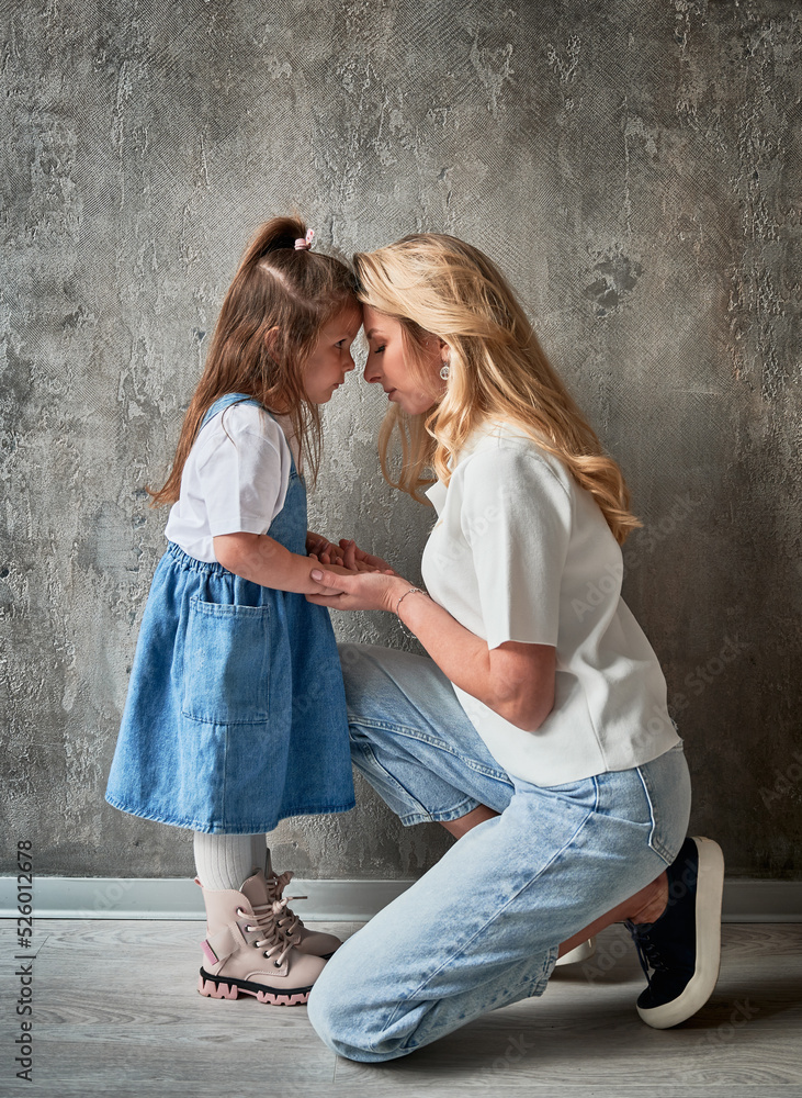Side view of cute child with mother standing against the wall at home. Blonde woman holding daughter hand. Family and motherhood concept. Side view of cute child with mother standing against the wall at home. Blonde woman holding daughter hand. Family and motherhood concept.