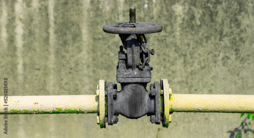 An old valve painted with thick black paint on a pipe, against the background of a wall.