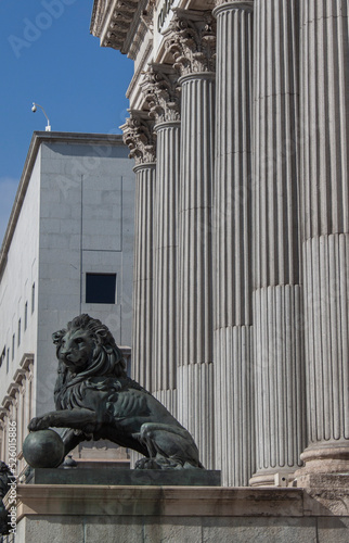 Bronze Lion statue at Congress of Deputies (Congreso de los Diputados) in Madrid, Spain, Europe. Historical columns building, gold entrance door in the background.