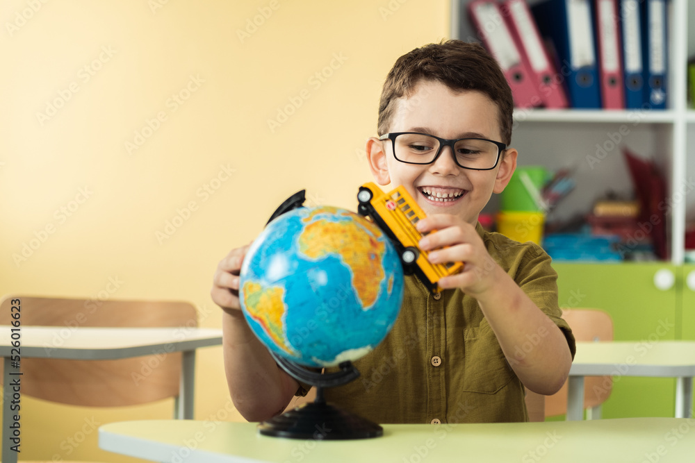 Cute caucasian schoolboy wearing glasses and sits at a desk in the classroom at elementary school. Little boy next the Globe playing with yellow school bus toy. Back to school concept Cute caucasian schoolboy wearing glasses and sits at a desk in the classroom at elementary school. Little boy next the Globe playing with yellow school bus toy. Back to school concept