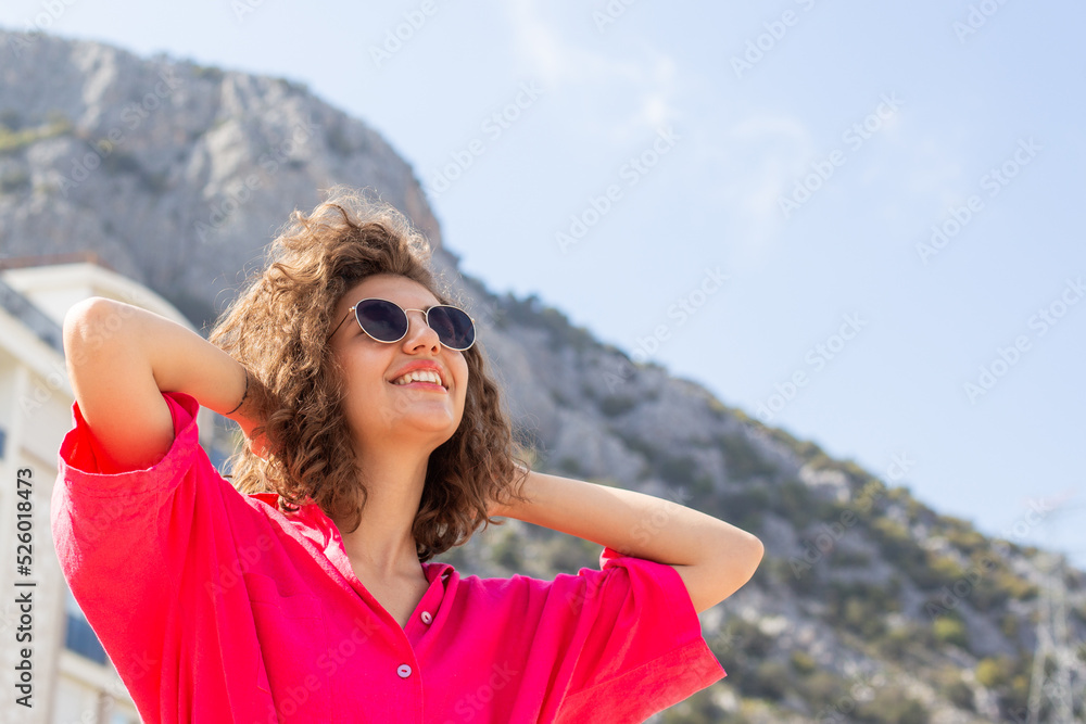young pretty curly haired woman in pink cloth and sunglasses having fun in the city with mountains background.