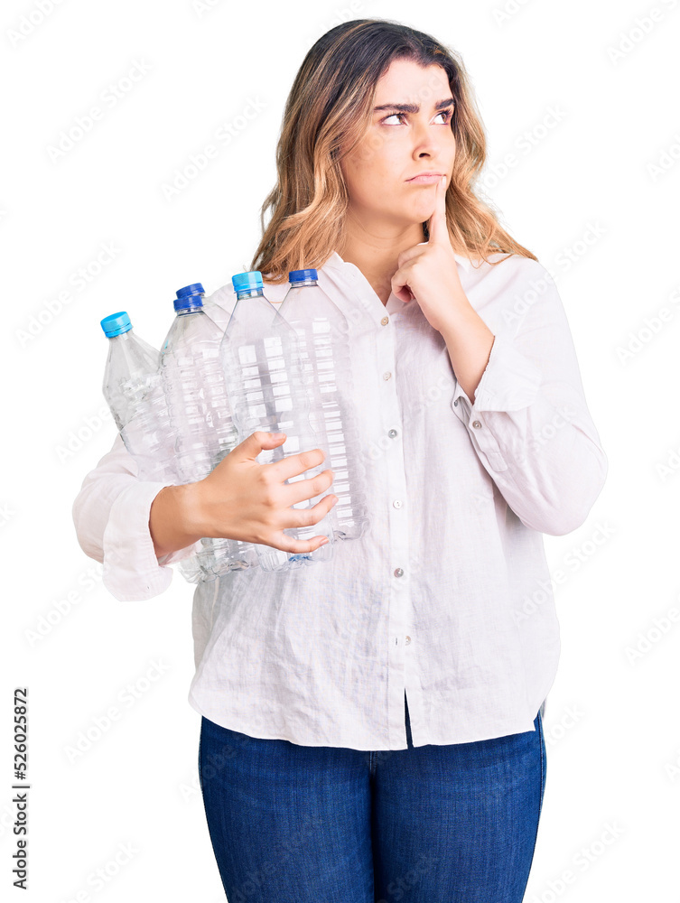 Young caucasian woman holding recycling plastic bottles serious face thinking about question with hand on chin, thoughtful about confusing idea Young caucasian woman holding recycling plastic bottles serious face thinking about question with hand on chin, thoughtful about confusing idea