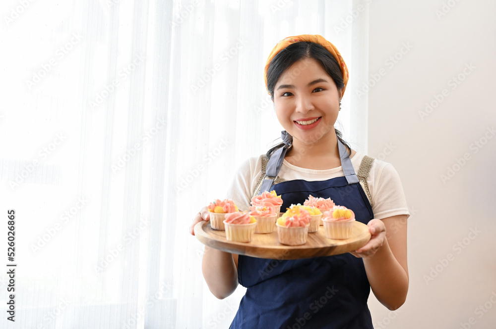 Beautiful Asian female baker in apron holding her finished delish cupcake on a wooden tray. Beautiful Asian female baker in apron holding her finished delish cupcake on a wooden tray.