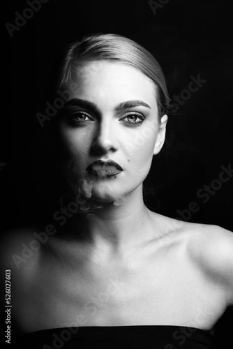 Fashion, style and make-up concept. Close-up black and white studio portrait of beautiful blonde woman blowing smoke from mouth. Model looking to camera with seductive look