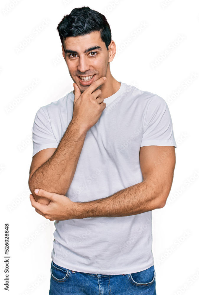 Handsome hispanic man wearing casual white t shirt looking confident at the camera with smile with crossed arms and hand raised on chin. thinking positive. Handsome hispanic man wearing casual white t shirt looking confident at the camera with smile with crossed arms and hand raised on chin. thinking positive.