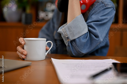 A hipster female in jean jacket at her working desk, holding a mug of coffee. cropped image
