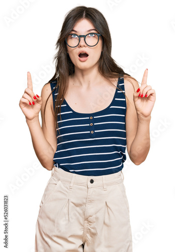 Young beautiful caucasian girl wearing casual clothes and glasses amazed and surprised looking up and pointing with fingers and raised arms.