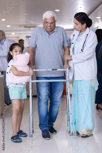 Granddaughter with female doctor teaching disabled grandfather to use walker at hospital
