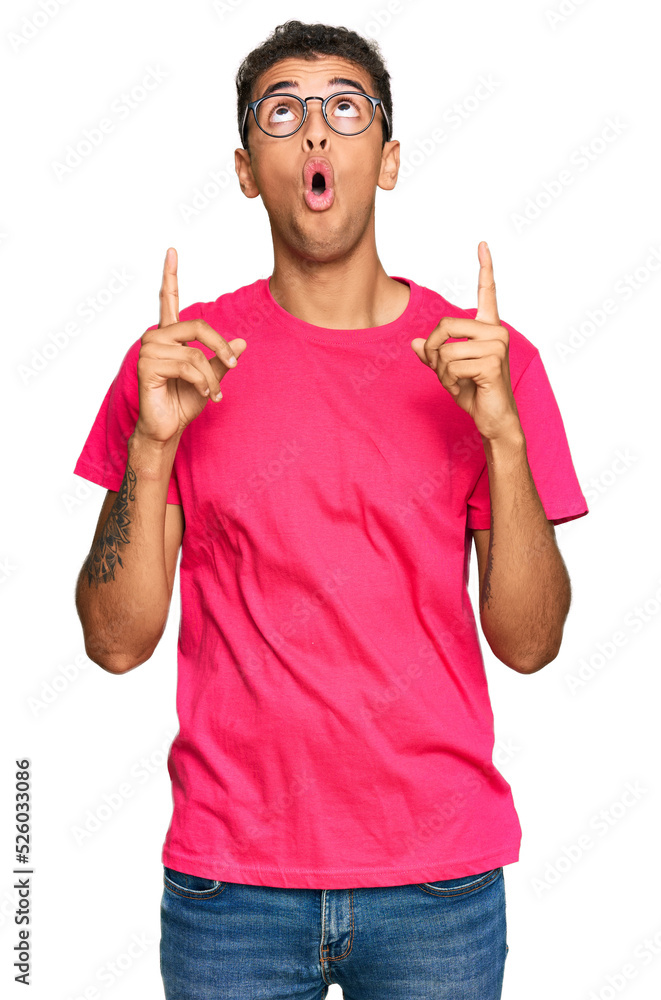 Young handsome african american man wearing casual clothes amazed and surprised looking up and pointing with fingers and raised arms. Young handsome african american man wearing casual clothes amazed and surprised looking up and pointing with fingers and raised arms.