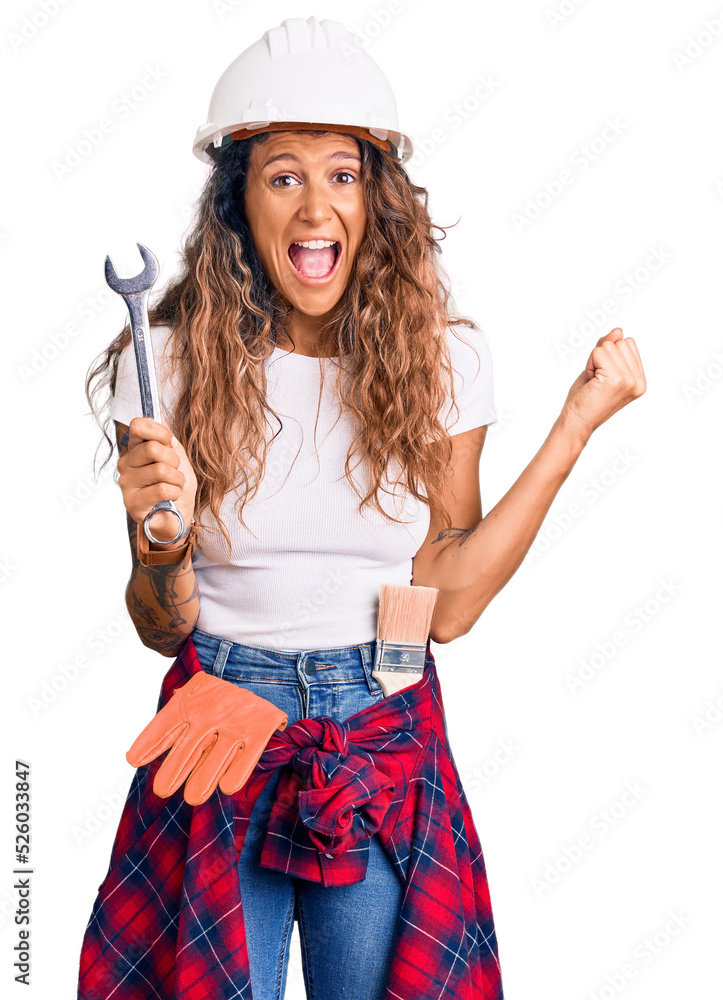 Young hispanic woman with tattoo wearing hardhat and builder clothes holding wrench screaming proud, celebrating victory and success very excited with raised arms Young hispanic woman with tattoo wearing hardhat and builder clothes holding wrench screaming proud, celebrating victory and success very excited with raised arms
