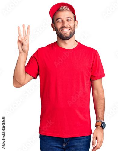 Young handsome blond man wearing t-shirt and cap showing and pointing up with fingers number three while smiling confident and happy.