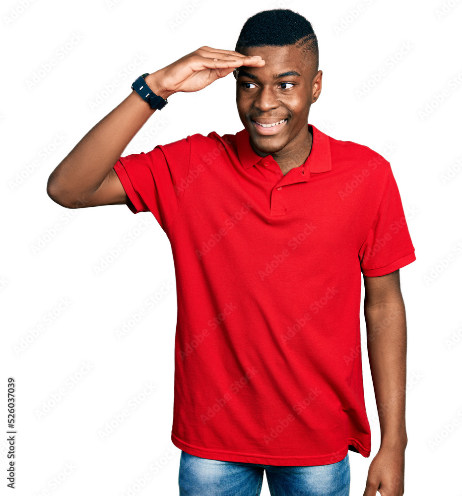 Young african american man wearing casual red t shirt very happy and smiling looking far away with hand over head. searching concept. Young african american man wearing casual red t shirt very happy and smiling looking far away with hand over head. searching concept.