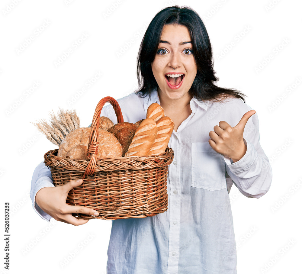Beautiful hispanic woman holding wicker basket with bread pointing thumb up to the side smiling happy with open mouth Beautiful hispanic woman holding wicker basket with bread pointing thumb up to the side smiling happy with open mouth