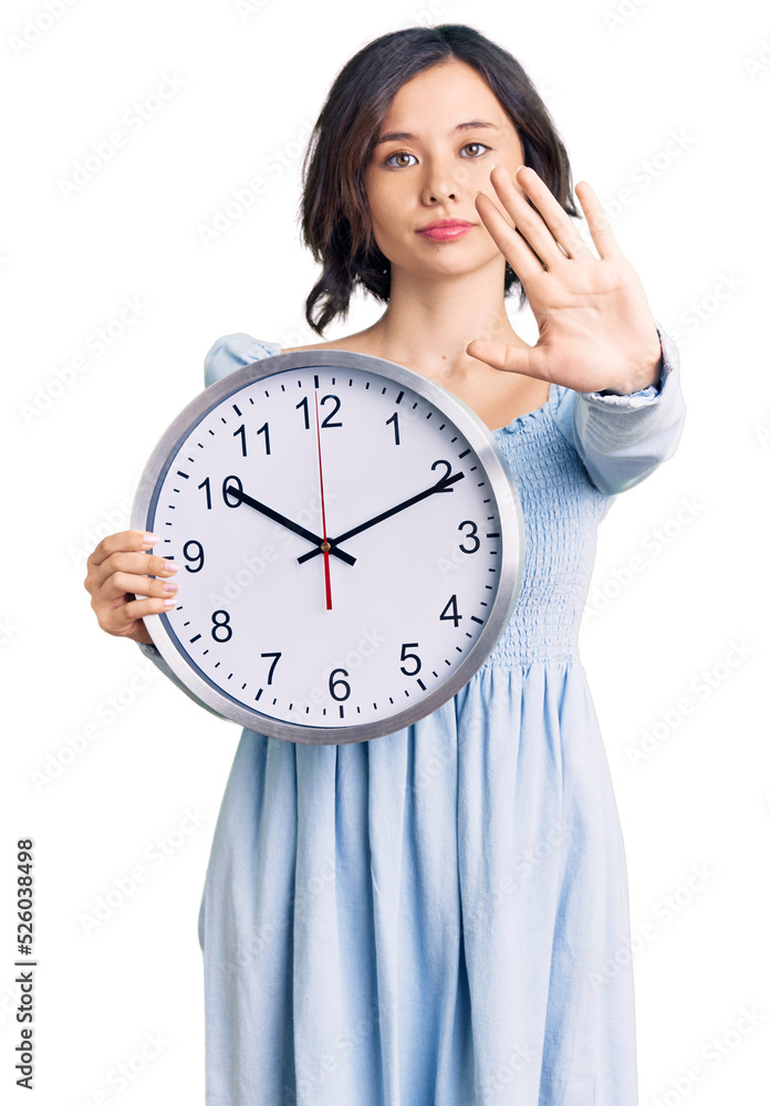 Young beautiful girl holding big clock with open hand doing stop sign with serious and confident expression, defense gesture Young beautiful girl holding big clock with open hand doing stop sign with serious and confident expression, defense gesture