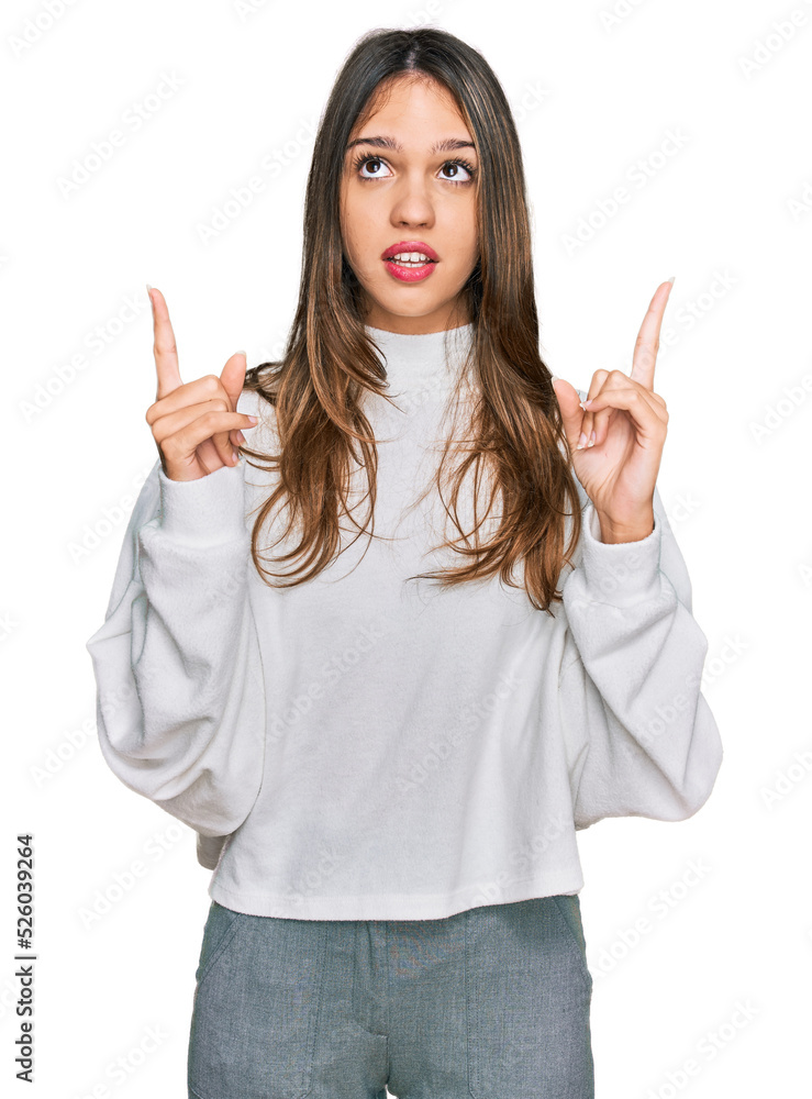 Young brunette woman wearing casual turtleneck sweater amazed and surprised looking up and pointing with fingers and raised arms. Young brunette woman wearing casual turtleneck sweater amazed and surprised looking up and pointing with fingers and raised arms.